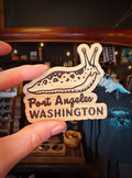 A hand holds a wooden souvenir magnet shaped like a snail with the text 'Port Angeles WASHINGTON' engraved on it, set against a blurred retail background.